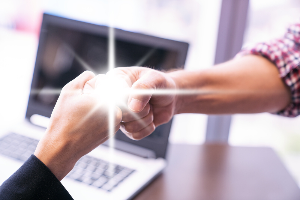 Two people fist-bumping in front of a laptop, with a glowing spark symbolizing trust and connection.